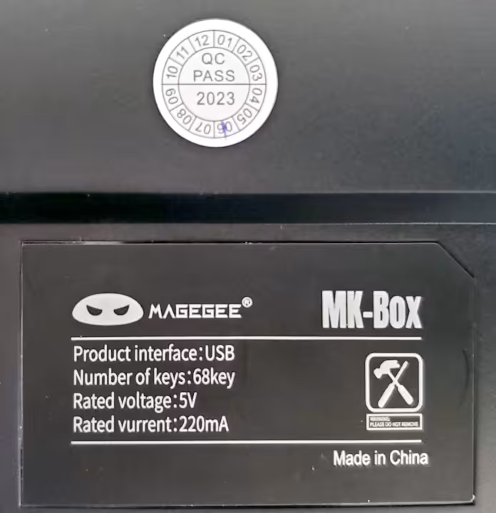 Closeup view of the back of a MageGee MK-Box keyboard showing the quality check circular sticker dated June 2023 and a brand label. The brand label displays made in China with make (MageGee), model (MK-Box) and general specifications of product interface (USB), number of keys (68), rated voltage (5V), and rated current (220mA).